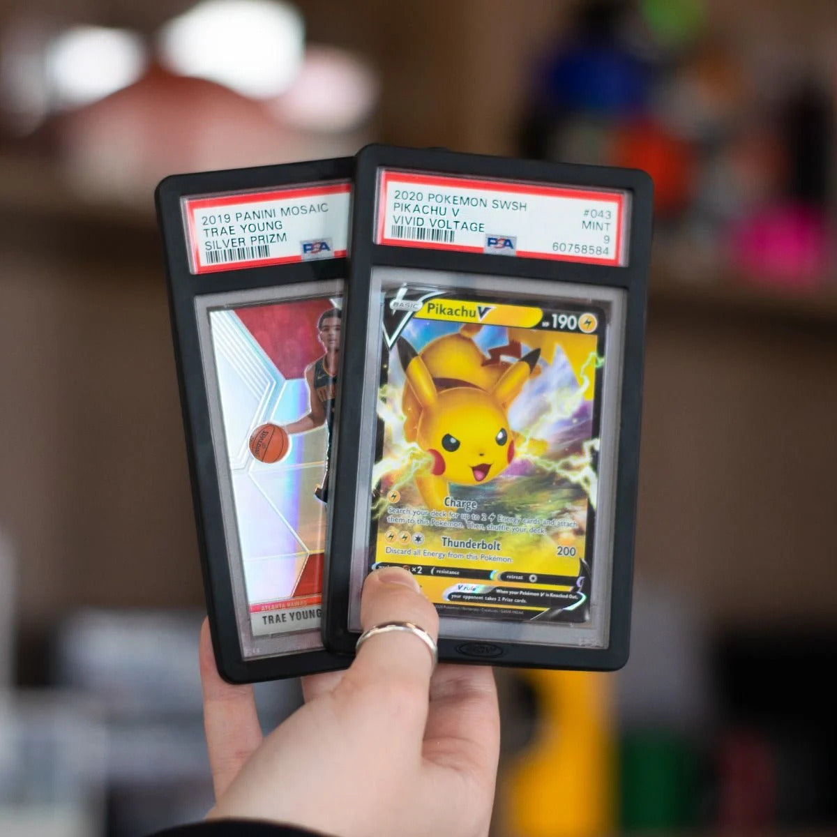 Two Black PSA Slab Case Bumpers in hand; one with 2019 Panini Mosaic Trae Young Silver Prizm PSA slab, other 2020 Pokémon SWSH Pikachu V Vivid Voltage PSA 9 slab