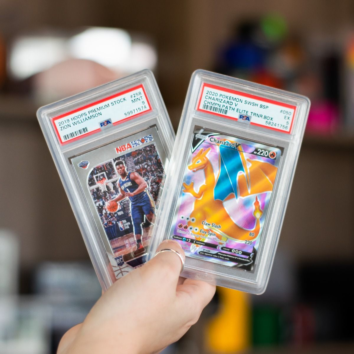 Two Clear PSA Slab Case Bumpers in hand; one with 2019 Hoops Premium Stock Zion Williamson RC #258  PSA 9 slab, other 2020 Pokémon SWSH BSP Charizard V Champion Path Elite Trainer Box PSA 5 slab